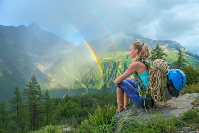 Young beautiful woman looking at a colorful double rainbow during summer rain in the mountains